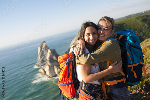 Happy mother and daughter hugging against seascape. Portrait of female backpackers posing in mountains in summer, looking at camera and smiling. Active family weekend concept