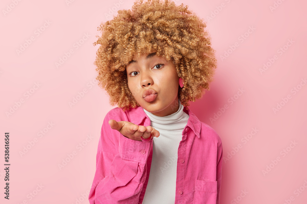 Cute romantic European woman blows air kiss sends mwah at camera has tender facial expression keeps lips rounded wears white comfortable turtleneck and shirt isolated over pink background adores you