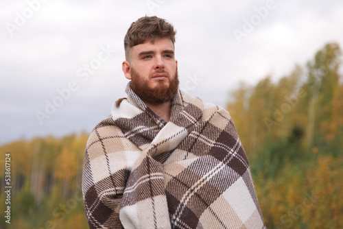 portrait of handsome bearded cozy guy, young serious pensive man with beard standing in golden autumn park or forest covering himself in blanket, plaid on natural background