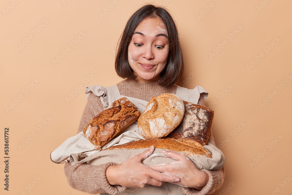 Happy dark haired Asian woman works in home bakery holds homemade yeast free bread stares at appetizing delicious food wears jumper and apron isolated over beige background. Healthy food concept Happy dark haired Asian woman works in home bakery holds homemade yeast free bread stares at appetizing delicious food wears jumper and apron isolated over beige background. Healthy food concept