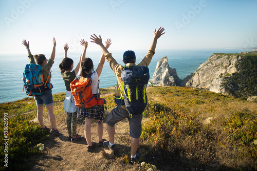 Excited family raising arms at precipice. Rear view of hiking parents with teen daughters enjoying beautiful view on mountain in summer. Active family weekend concept