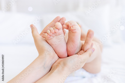 mom's hands hold the baby's feet in focus on a white crib at home with white cotton bedding, the concept of baby goods and accessories and mom's love and care