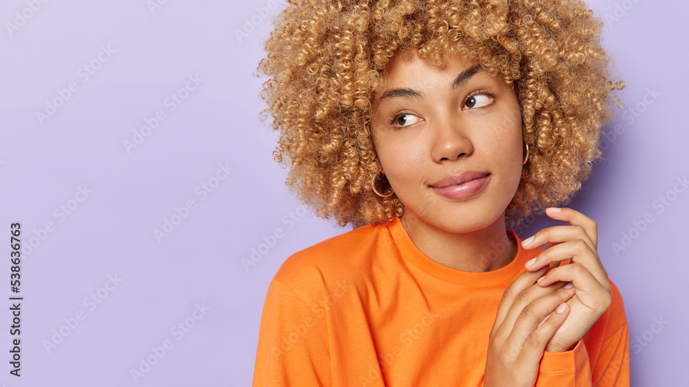 Studio shot of good looking curly haired woman keeps hands together looks aside pensively wears casual orange jumper isolated over purple background blank space for your promotional content. Studio shot of good looking curly haired woman keeps hands together looks aside pensively wears casual orange jumper isolated over purple background blank space for your promotional content.