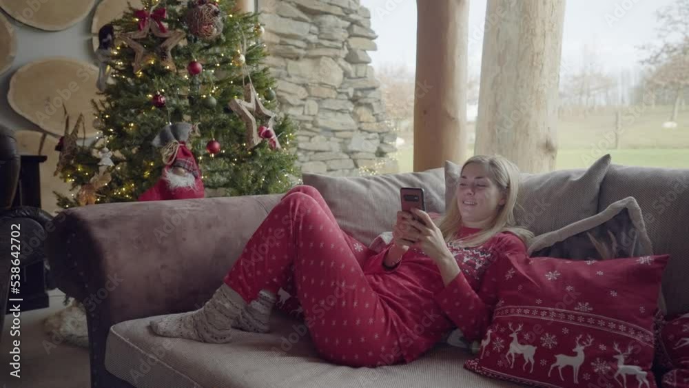 Full shot - Shot of a blonde woman chilling on a sofa, dressed in red pyjama and Christmas socks, surrounded by Christmas decorations, on a calm winter morning Full shot - Shot of a blonde woman chilling on a sofa, dressed in red pyjama and Christmas socks, surrounded by Christmas decorations, on a calm winter morning