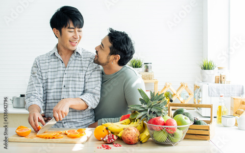 Gay LGBT sweet Asian couple wearing pajamas  smiling  looking each other  hugging with happiness and love while making breakfast with healthy fruits in kitchen at home in morning. Lifestyle Concept.