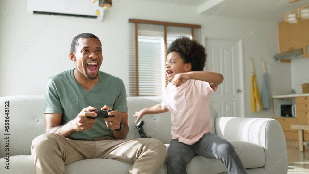Cheerful Black African American Father and his Little son in Afro hair sitting on sofa using joysticks or game controllers playing console video games at home Cheerful Black African American Father and his Little son in Afro hair sitting on sofa using joysticks or game controllers playing console video games at home