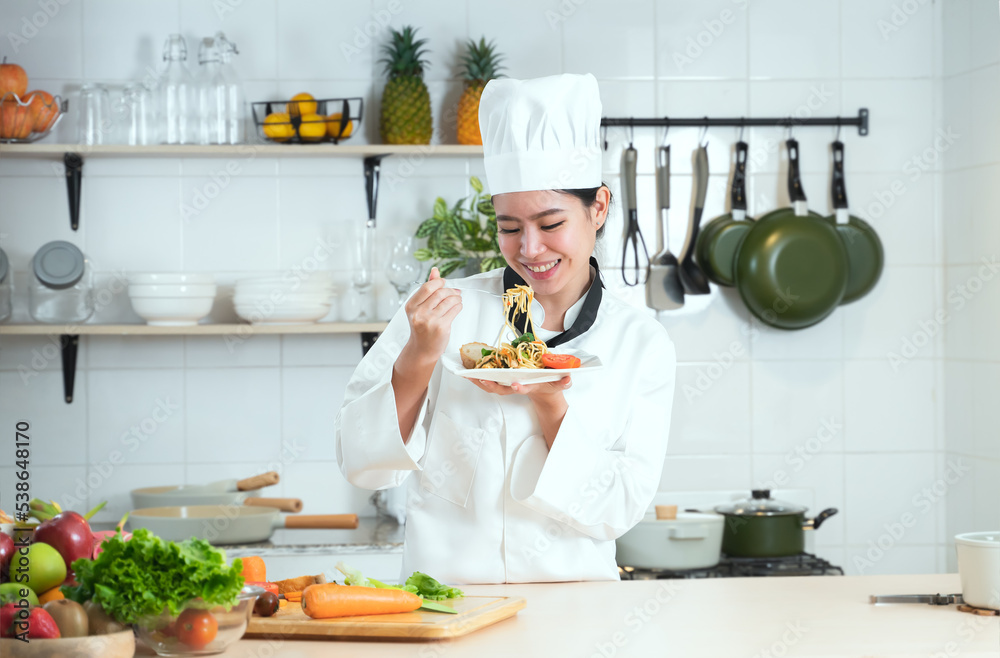 Portrait of Asian young beautiful chef woman wear white chef uniform and hat, smiling looking at spaghetti dish with delicious face, working at restaurant kitchen, fruits and vegetables on table Portrait of Asian young beautiful chef woman wear white chef uniform and hat, smiling looking at spaghetti dish with delicious face, working at restaurant kitchen, fruits and vegetables on table