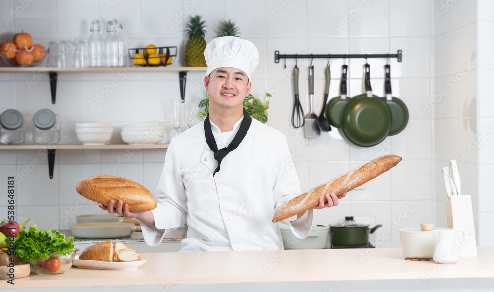 Portrait of Asian young handsome chef man wear white chef uniform and hat, holding loaf of handmade french bread in hands, smiling looking at camera, working at restaurant kitchen Portrait of Asian young handsome chef man wear white chef uniform and hat, holding loaf of handmade french bread in hands, smiling looking at camera, working at restaurant kitchen