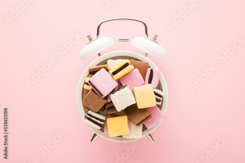 Colorful jelly gummy candies on white alarm clock on light pink table background. Pastel color. Sweets time concept. Closeup. Top down view.