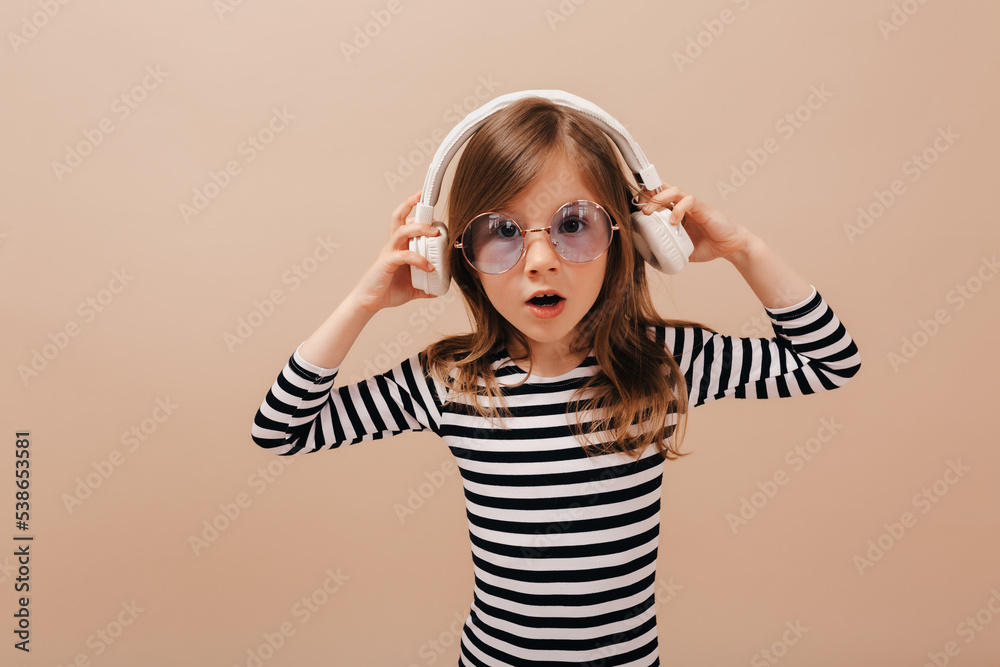 Charming good-humored little girl in striped shirt and round glasses inn headphones posing at camera with surprised emotions over beige background Charming good-humored little girl in striped shirt and round glasses inn headphones posing at camera with surprised emotions over beige background