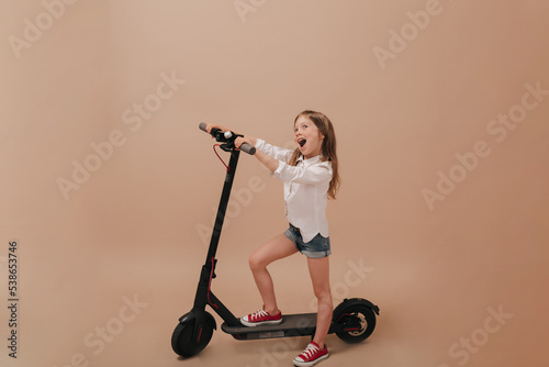 Full-lenght photo of excited charming little girl wearing white shirt and shorts playing and having fun with electro scooter over beige background. 