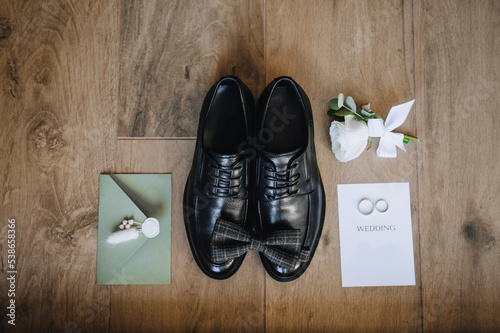 Groom's black shoes, bow tie, boutonniere, gold rings, an envelope with a heart stamp, a sheet of paper lie on a wooden background. Wedding photo of accessories and details, top view.