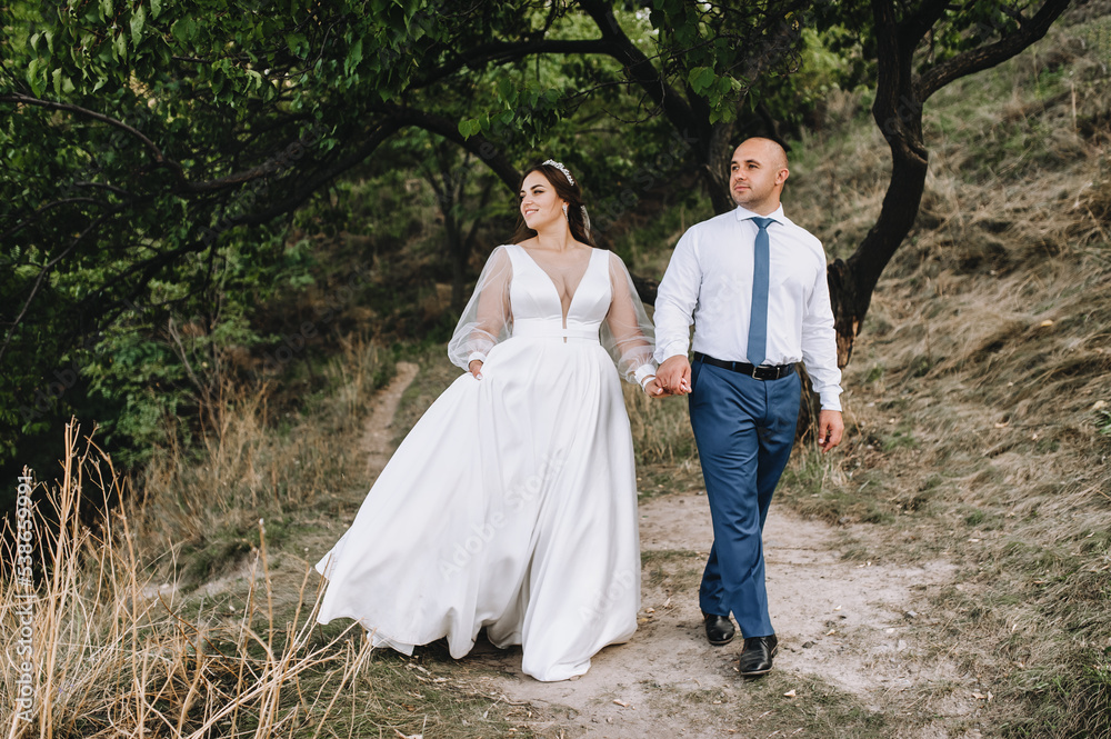 A stylish, loving groom and a beautiful, young, smiling bride in a white dress are walking in a park in nature, holding hands. Wedding photography, portrait. A stylish, loving groom and a beautiful, young, smiling bride in a white dress are walking in a park in nature, holding hands. Wedding photography, portrait.