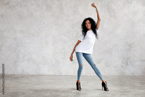 Beautiful African American woman dances  moves and smiles on light gray minimalist background. Young model wears jeans  high heels and white T-shirt