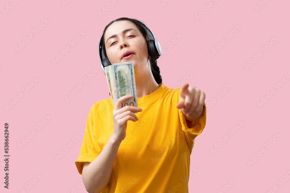 Winning the lottery jackpot. A young Caucasian happy woman in headphones sing a bundle of dollars as a microphone. Pink background. The concept of successful business and investment Winning the lottery jackpot. A young Caucasian happy woman in headphones sing a bundle of dollars as a microphone. Pink background. The concept of successful business and investment