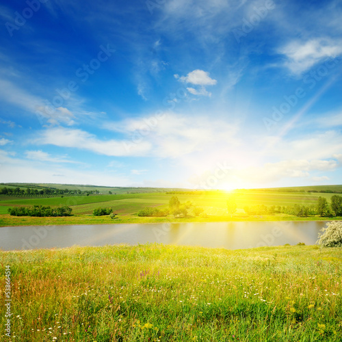 Meadow, lake, agricultural fields and sunrise on blue sky.