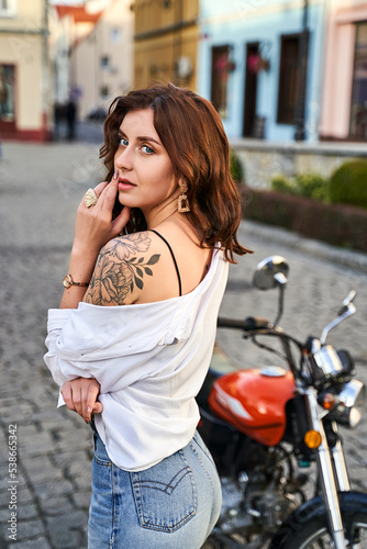 Obraz na płótnie Young beautiful woman shows her bare shoulder with a tattoo standing by a bike on the street w salonie