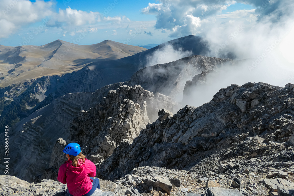 Rear view of woman with climbing helmet sitting on cloud covered mountain summit of Mytikas Mount Olympus, Mt Olympus National Park, Macedonia, Greece, Europe. View of rocky ridges and mountain ranges