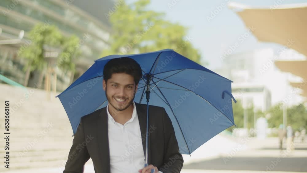 Young muslim man opening blue umbrella, hiding from sun, posing and smiling at camera. Arabic guy in suit looking at umbrella, spinning it. City background. Fashion, lifestyle concept.