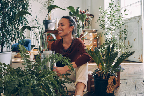 Fotografia Happy young woman watering houseplants while sitting on the floor at home