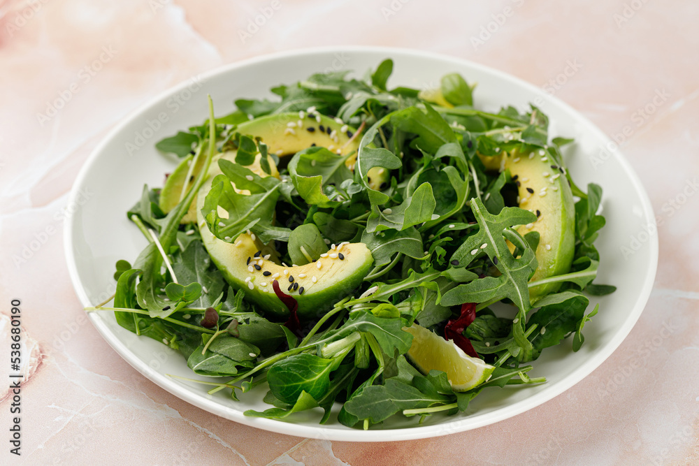 Vegan green salad with avocado and mix lettuce, arugula on white plate and beige marble background.