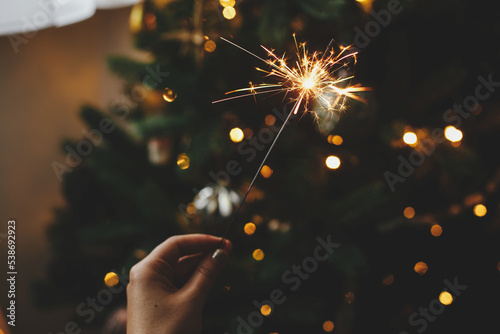 Burning sparkler in female hand on background of christmas tree lights in dark room. Happy New Year! Hand holding firework against stylish decorated tree with illumination. Atmospheric time