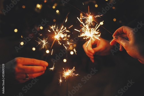Happy New Year! Friends celebrating with burning sparklers in hands against christmas tree lights in dark room. Hands holding fireworks on background of stylish decorated illuminated tree. Moody