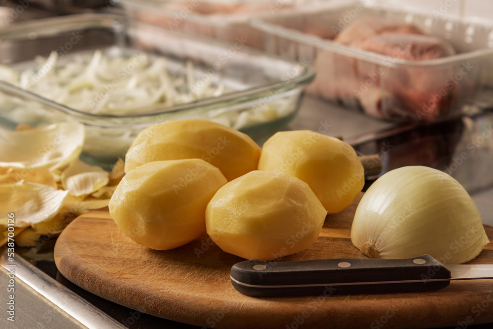 Peeled potatoes on a cutting board and a knife, in the background some trays with onions and out of focus chicken thighs, in the kitchen of the home. Peeled potatoes on a cutting board and a knife, in the background some trays with onions and out of focus chicken thighs, in the kitchen of the home.