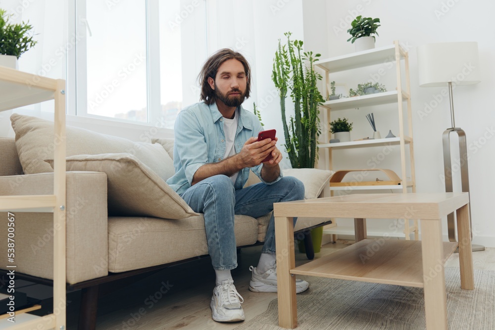 A freelance man in a white T-shirt, blue jeans, and shirt sits on the couch with his phone at home on his day off and plays games
