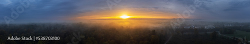 Panoramic view over a forest and a city on a foggy autumn morning while the sun rises in the background