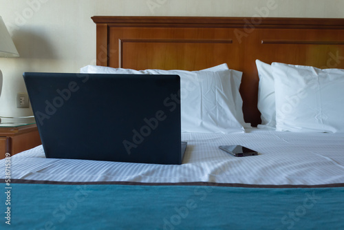 Laptop and cell phone on a bed with pillows and white sheets.
