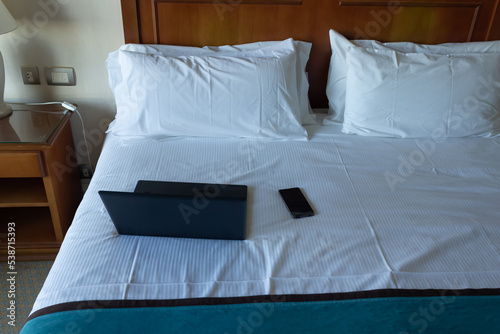 Laptop and cell phone on a bed with pillows and white sheets.