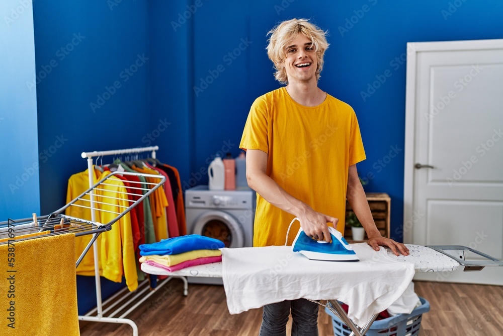 Young blond man smiling confident ironing clothes at laundry room