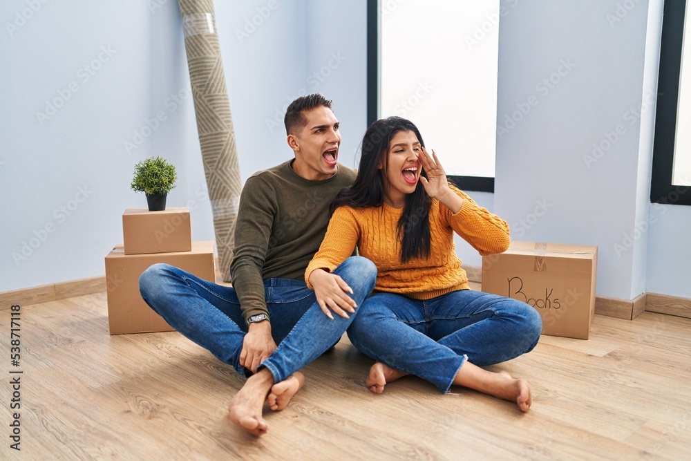 Young couple sitting on the floor at new home shouting and screaming loud to side with hand on mouth. communication concept. Young couple sitting on the floor at new home shouting and screaming loud to side with hand on mouth. communication concept.