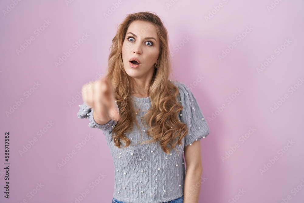 Beautiful blonde woman standing over pink background pointing with finger surprised ahead, open mouth amazed expression, something on the front Beautiful blonde woman standing over pink background pointing with finger surprised ahead, open mouth amazed expression, something on the front