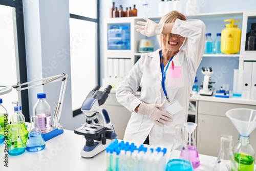 Middle age blonde woman working at scientist laboratory covering eyes with arm smiling cheerful and funny. blind concept.