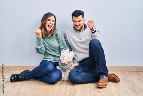 Young hispanic couple sitting on the floor with dog very happy and excited doing winner gesture with arms raised, smiling and screaming for success. celebration concept.