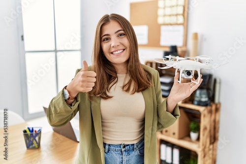 Young brunette woman holding drone at architect office smiling happy and positive, thumb up doing excellent and approval sign