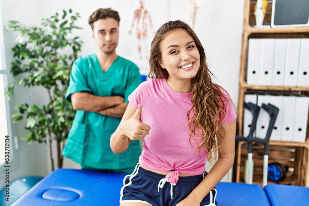 Young hispanic woman at physiotherapist appointment doing happy thumbs up gesture with hand. approving expression looking at the camera showing success.