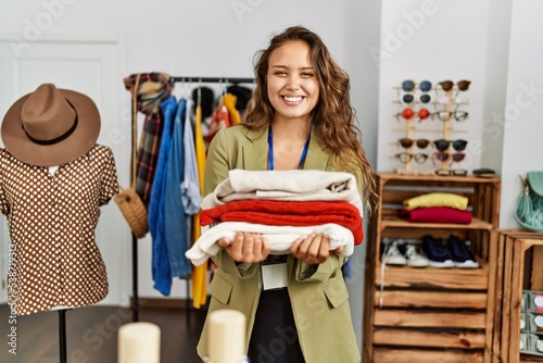 Young hispanic shopkeeper woman smiling happy holding stack of sweater at clothing store.