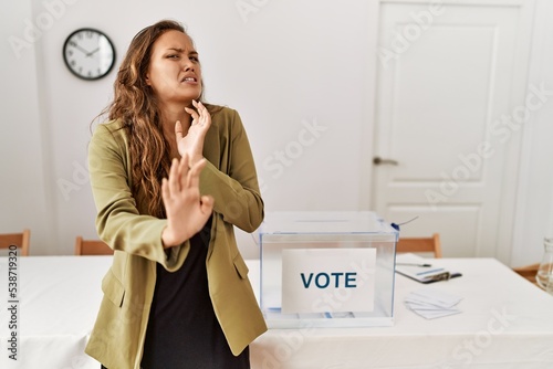 Beautiful hispanic woman standing at political campaign room disgusted expression, displeased and fearful doing disgust face because aversion reaction. with hands raised