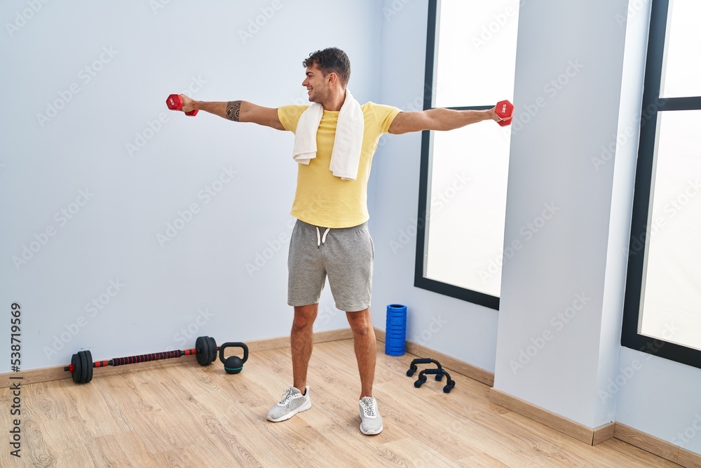 Young hispanic man smiling confident using dumbbells training at sport center