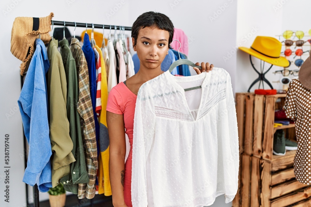 Young hispanic woman with short hair shopping at retail boutique thinking attitude and sober expression looking self confident
