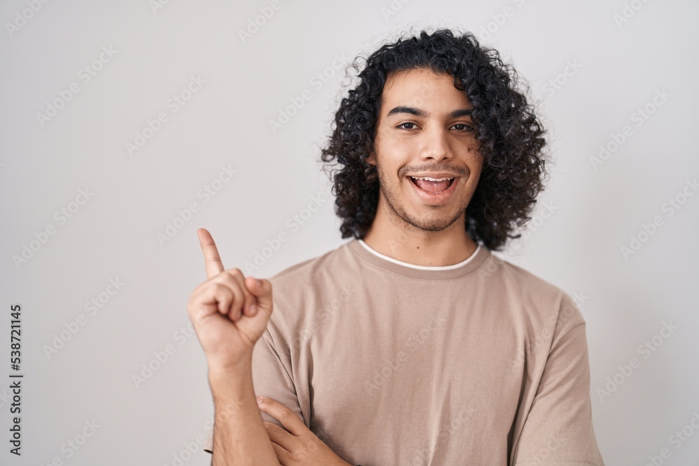 Hispanic man with curly hair standing over white background with a big smile on face, pointing with hand finger to the side looking at the camera. Hispanic man with curly hair standing over white background with a big smile on face, pointing with hand finger to the side looking at the camera.