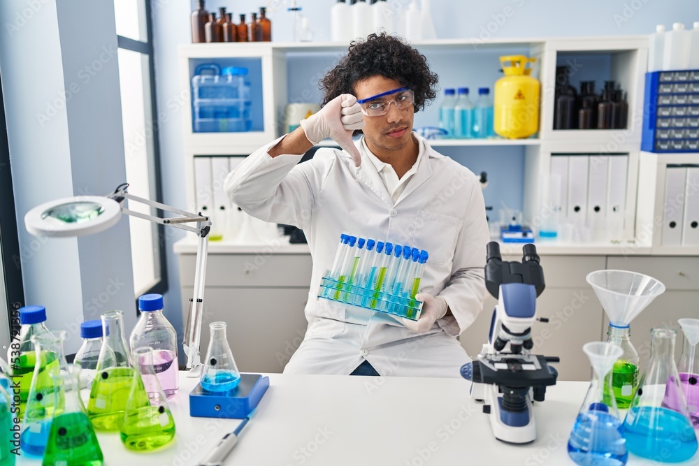 Hispanic man with curly hair working at scientist laboratory with angry face, negative sign showing dislike with thumbs down, rejection concept Hispanic man with curly hair working at scientist laboratory with angry face, negative sign showing dislike with thumbs down, rejection concept