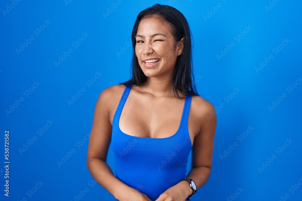 Hispanic woman standing over blue background winking looking at the camera with sexy expression, cheerful and happy face. Hispanic woman standing over blue background winking looking at the camera with sexy expression, cheerful and happy face.