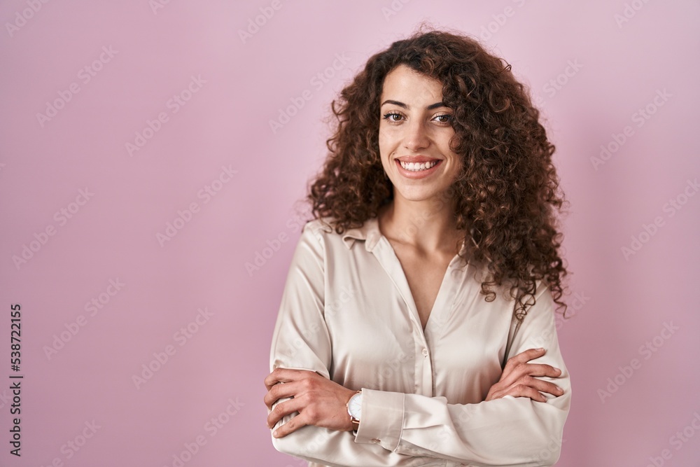 Hispanic woman with curly hair standing over pink background happy face smiling with crossed arms looking at the camera. positive person. Hispanic woman with curly hair standing over pink background happy face smiling with crossed arms looking at the camera. positive person.