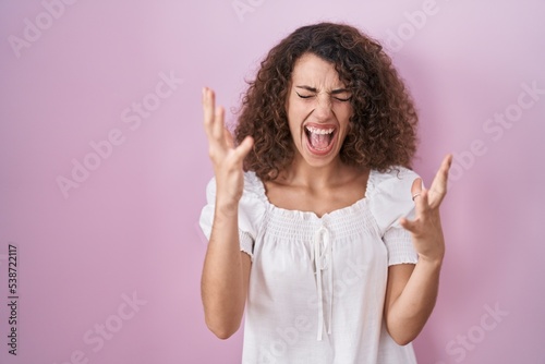 Hispanic woman with curly hair standing over pink background celebrating mad and crazy for success with arms raised and closed eyes screaming excited. winner concept