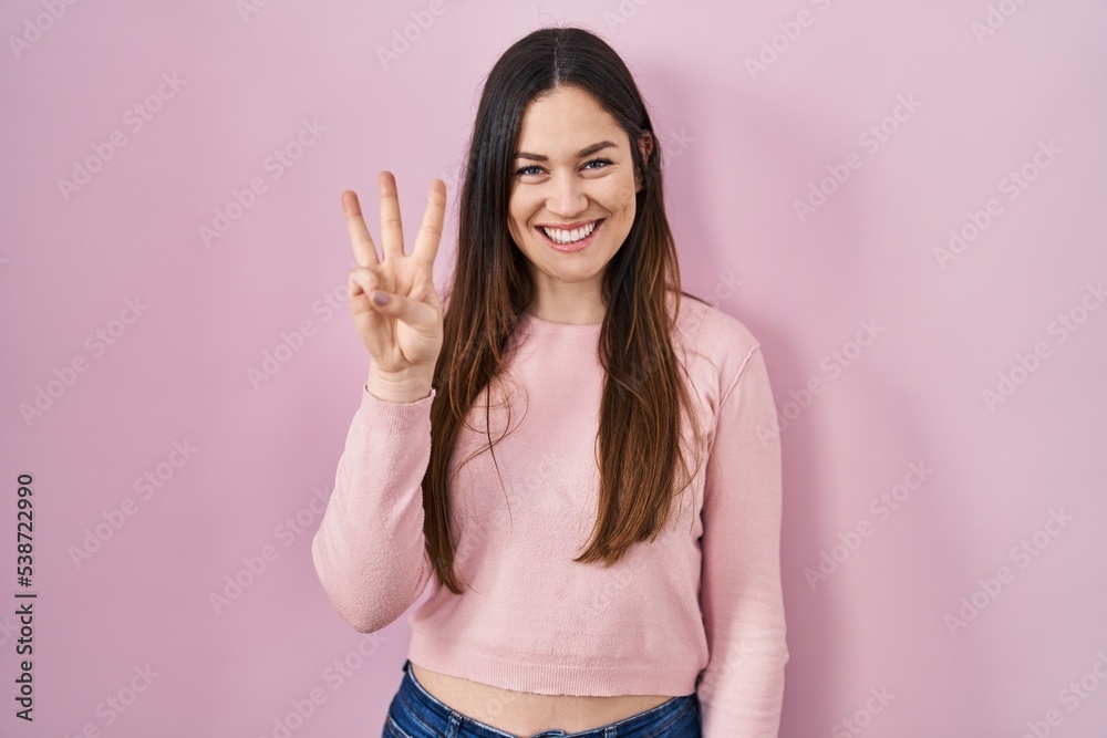 Young brunette woman standing over pink background showing and pointing up with fingers number three while smiling confident and happy. Young brunette woman standing over pink background showing and pointing up with fingers number three while smiling confident and happy.