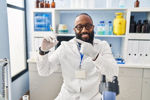 African american man working at scientist laboratory holding syringe smiling happy pointing with hand and finger